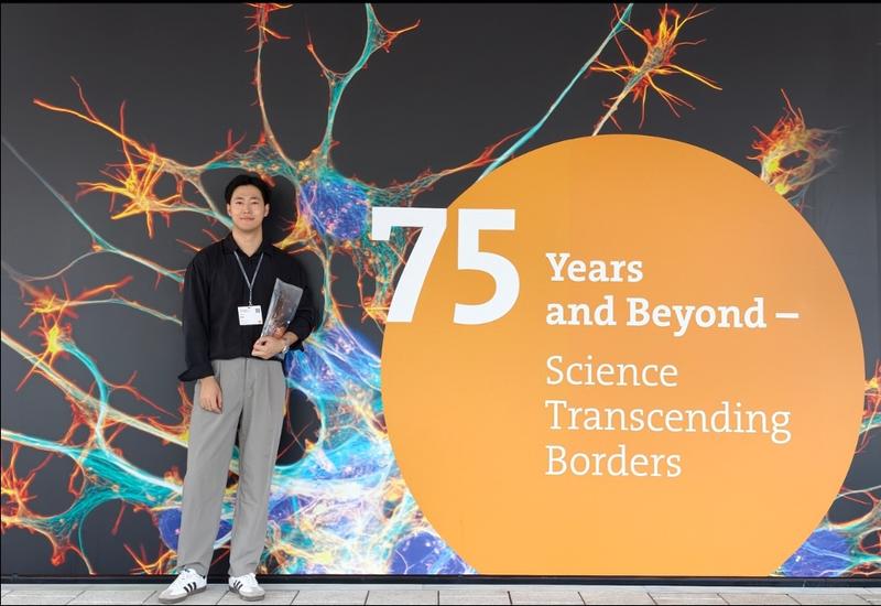 A young man stands in front of a colorful neuron-themed display at the Lindau Nobel Laureate Meetings. Beside him, a large orange circle reads, '75 Years and Beyond – Science Transcending Borders.'