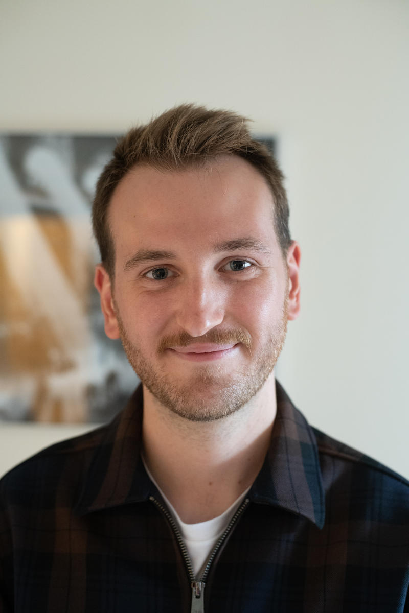 A close-up portrait of a smiling man with short brown hair and a light beard, wearing a dark plaid shirt over a white T-shirt, standing indoors against a softly blurred background.