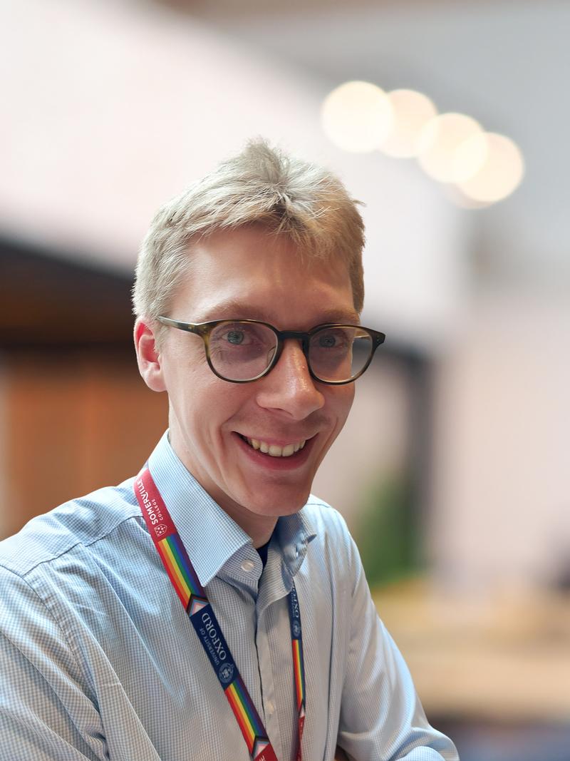 A smiling person with glasses and short blond hair in a light blue shirt, wearing an Oxford lanyard, poses indoors with a softly blurred background.