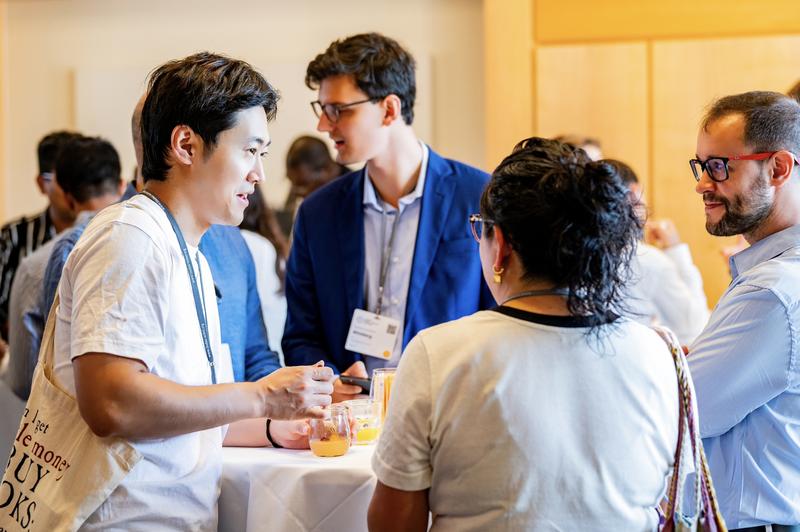 Attendees converse around a standing table during a networking reception, with drinks in hand and other guests mingling in the warmly lit background.