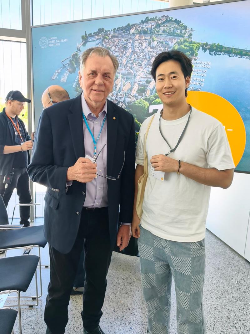 Two conference attendees pose together in front of a large Lindau Nobel Laureate Meetings display. The older person on the left wears a dark suit jacket, the younger person on the right wears a white T-shirt and patterned trousers.