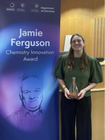 A smiling woman holds a trophy beside a banner for the Jamie Ferguson Chemistry Innovation Award at the University of Oxford Department of Chemistry.