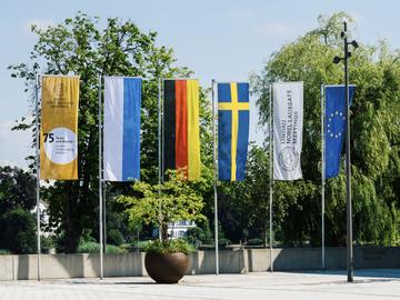 Six outdoor flags stand beside trees, including Lindau Nobel Laureate Meetings banners and the flags of Bavaria, Germany, Sweden, and the European Union.