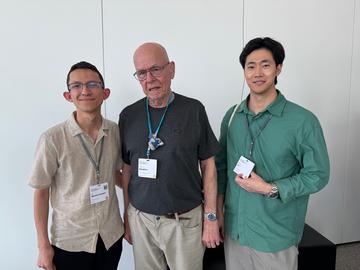 Three conference attendees stand side by side against a white wall, smiling. The person on the left wears a beige short-sleeve shirt and glasses, the person in the center wears a dark T-shirt and glasses, the person on the right wears a green shirt.
